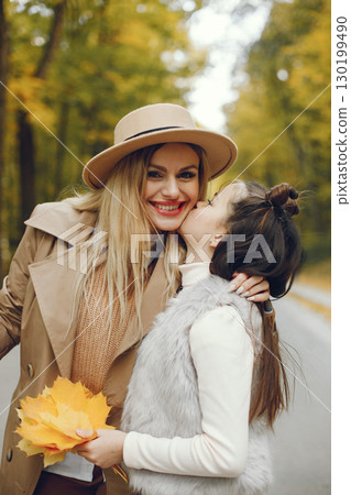 Woman and her little girl walking in the autumn park. Mother and daughter posing for a photo with an autumn leaves. Woman wearing beige coat and a hat. 130199490