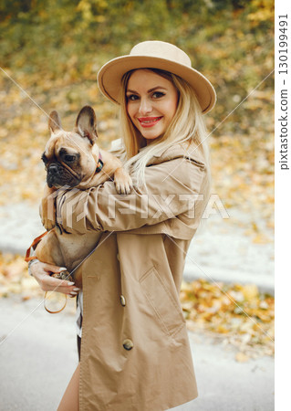 Woman and dog play and have fun in the autumn park. Brown french bulldog with female owner spend a day at the park walking. Woman wearing beige coat and a hat. 130199491