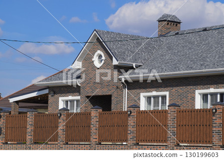 Asphalt shingle. Decorative bitumen shingles on the roof of a brick house. Fence made of corrugated metal. Asphalt shingle. Decorative bitumen shingles on the roof of a brick house. Fence made of corrugated metal. 130199603