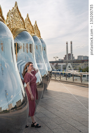 Woman in a rose dress poses by giant reflective matryoshka sculptures with ornate golden tops on Taras Shevchenko Embankment in Moscow Woman in a rose dress poses by giant reflective matryoshka sculptures with ornate golden tops on Taras Shevchenko Embankment in Moscow 130200785