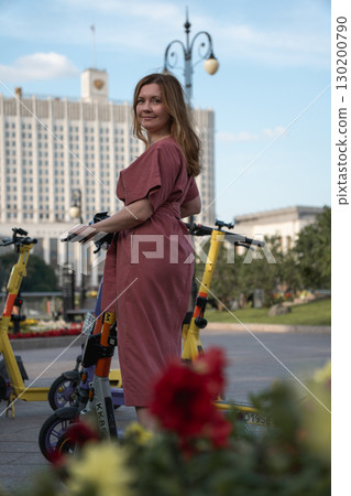 Smiling woman in a rose dress standing with an electric scooter on Taras Shevchenko Embankment, Moscow, with the White House in the background Smiling woman in a rose dress standing with an electric scooter on Taras Shevchenko Embankment, Moscow, with the White House in the background 130200790