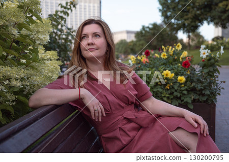 Woman in a rose dress sits on a bench near blooming hydrangeas, gazing thoughtfully into the distance with a serene expression 130200795