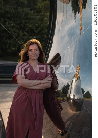 Woman in rose dress stands with arms crossed by mirrored matryoshka sculpture, evening sunlight highlighting her hair 130200801
