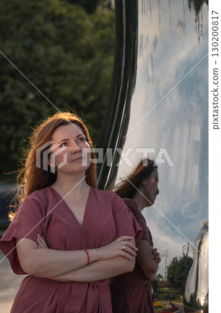 Woman in rose dress stands with arms crossed by mirrored matryoshka sculpture, evening sunlight highlighting her hair 130200817