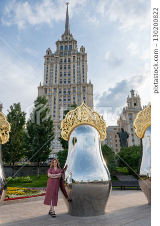 Woman in rose dress poses by mirrored matryoshka sculpture with golden ornament against the backdrop of the historic Radisson Royal Hotel Woman in rose dress poses by mirrored matryoshka sculpture with golden ornament against the backdrop of the historic Radisson Royal Hotel 130200822