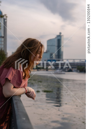 Woman in a red dress leaning on a riverside railing, looking down thoughtfully with city buildings and soft sunset light in the background Woman in a red dress leaning on a riverside railing, looking down thoughtfully with city buildings and soft sunset light in the background 130200844