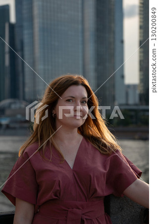 Portrait of a woman in a red dress with loose hair, softly lit by evening sunlight, standing against a background of modern skyscrapers 130200849