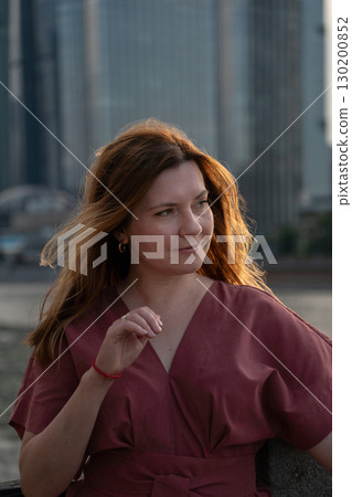 Woman in a red dress with loose hair looks thoughtfully to the side, standing outdoors with blurred modern skyscrapers in the background 130200852