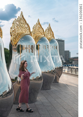Woman in a rose dress poses by giant reflective matryoshka sculptures with ornate golden tops on Taras Shevchenko Embankment in Moscow 130200861