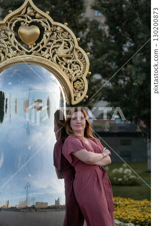 Woman in a rose dress leans against a giant reflective matryoshka sculpture with ornate golden crown on Taras Shevchenko Embankment in Moscow Woman in a rose dress leans against a giant reflective matryoshka sculpture with ornate golden crown on Taras Shevchenko Embankment in Moscow 130200873