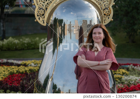 Woman in a rose dress leans against a giant reflective matryoshka sculpture with ornate golden crown on Taras Shevchenko Embankment in Moscow 130200874