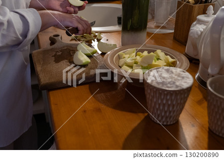 A person slices green apples on a wooden cutting board next to a bowl of chopped apples and a cup of flour on the kitchen table 130200890