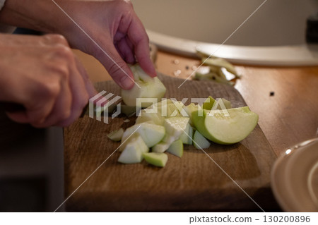 A person slices green apples on a wooden cutting board next to a bowl of chopped apples and a cup of flour on the kitchen table A person slices green apples on a wooden cutting board next to a bowl of chopped apples and a cup of flour on the kitchen table 130200896