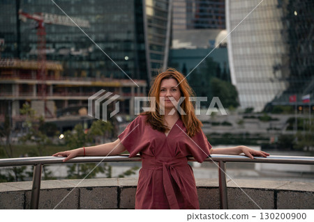 Woman in a burgundy dress poses against the backdrop of the modern skyscrapers of Moscow City 130200900