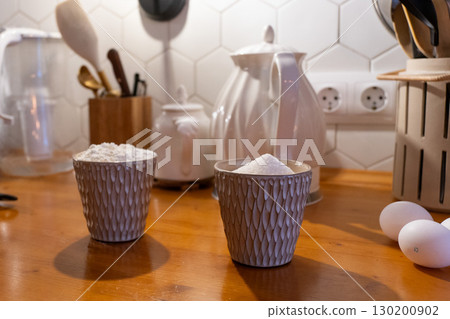 Ceramic cups with flour and sugar stand on a wooden kitchen counter, alongside eggs and a teapot, creating a homely cooking atmosphere 130200902
