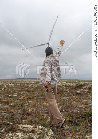 Person in a gray jacket and beanie reaches up towards a wind turbine in the tundra of Teriberka, Russia 130200905