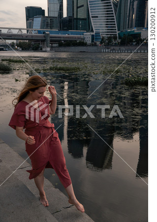Woman in a burgundy dress walks barefoot by the Moscow River,balancing with arms outstretched against the backdrop of water and skyscraper reflections Woman in a burgundy dress walks barefoot by the Moscow River,balancing with arms outstretched against the backdrop of water and skyscraper reflections 130200912