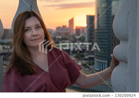 Woman in a rose dress standing on a rooftop terrace at sunset with wind in her hair, framed by ornate balustrades Woman in a rose dress standing on a rooftop terrace at sunset with wind in her hair, framed by ornate balustrades 130200924