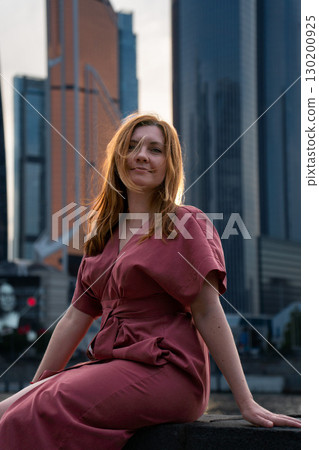 Woman in a red dress sits by the river with skyscrapers of Moscow City in the background, softly lit by sunset Woman in a red dress sits by the river with skyscrapers of Moscow City in the background, softly lit by sunset 130200925