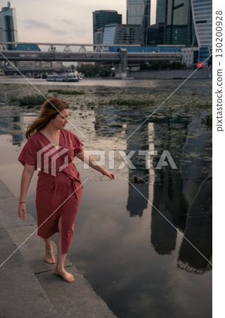 Woman in a burgundy dress walks barefoot by the Moscow River,balancing with arms outstretched against the backdrop of water and skyscraper reflections 130200928