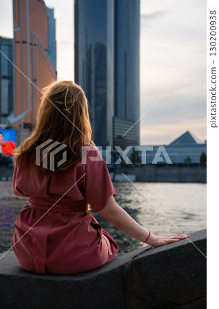 Woman in a burgundy dress sits on a stone embankment by the Moscow River, facing the skyscrapers of Moscow International Business Center at sunset Woman in a burgundy dress sits on a stone embankment by the Moscow River, facing the skyscrapers of Moscow International Business Center at sunset 130200938