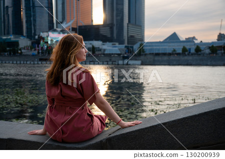 Woman in a burgundy dress sits on the Moscow River embankment at sunset, with skyscrapers of Moscow City in the background Woman in a burgundy dress sits on the Moscow River embankment at sunset, with skyscrapers of Moscow City in the background 130200939