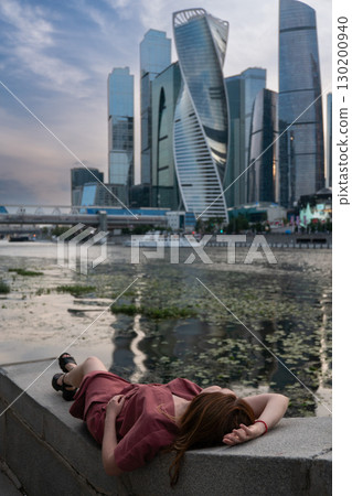 Young woman in a burgundy dress relaxing on a stone embankment, lying with her head back and hand resting on her forehead near the water Young woman in a burgundy dress relaxing on a stone embankment, lying with her head back and hand resting on her forehead near the water 130200940