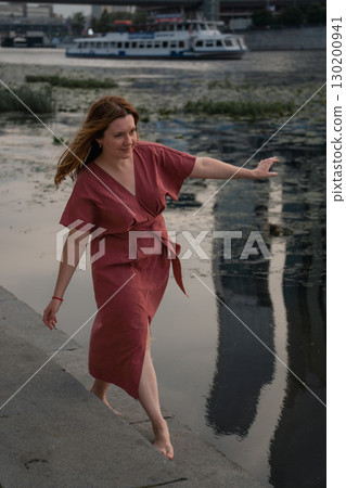 Woman in a burgundy dress walks barefoot by the Moscow River,balancing with arms outstretched against the backdrop of water and skyscraper reflections 130200941