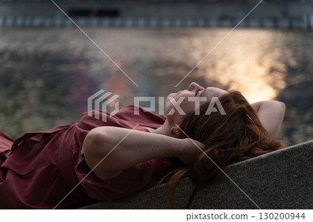 Young woman with red hair in a burgundy dress relaxing by the riverbank at sunset, with warm golden reflections shimmering on the water Young woman with red hair in a burgundy dress relaxing by the riverbank at sunset, with warm golden reflections shimmering on the water 130200944