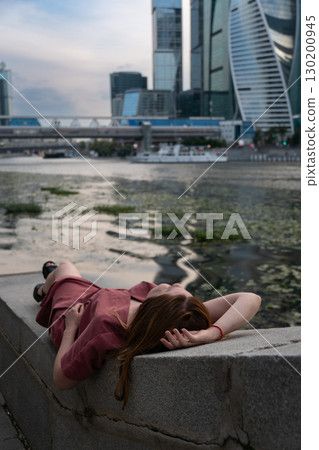 Young woman in a burgundy dress relaxing on a stone embankment, lying with her head back and hand resting on her forehead near the water Young woman in a burgundy dress relaxing on a stone embankment, lying with her head back and hand resting on her forehead near the water 130200945