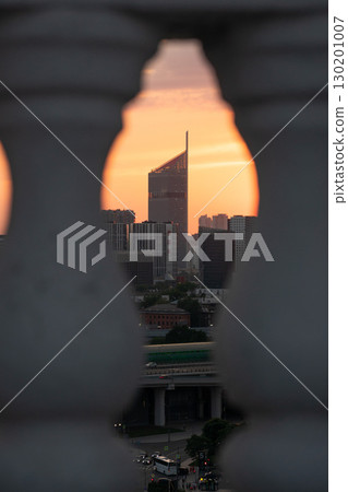 Modern skyscraper framed between blurred architectural balusters at sunset, with a warm orange sky casting a glow over the urban skyline Modern skyscraper framed between blurred architectural balusters at sunset, with a warm orange sky casting a glow over the urban skyline 130201007