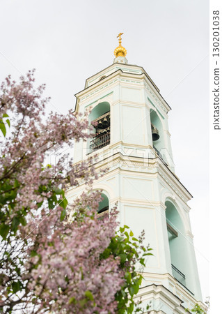 The bell tower of the Church of Sergius of Radonezh in Rogozhskaya Sloboda rises behind blooming lilac branches. Heritage preservation, religious tourism, and the seasonal beauty of urban architecture The bell tower of the Church of Sergius of Radonezh in Rogozhskaya Sloboda rises behind blooming lilac branches. Heritage preservation, religious tourism, and the seasonal beauty of urban architecture 130201038
