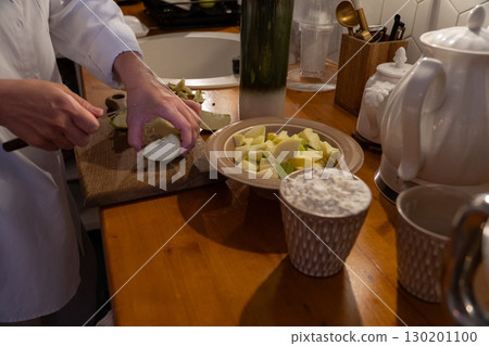A person slices green apples on a wooden cutting board next to a bowl of chopped apples and a cup of flour on the kitchen table 130201100