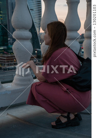 Woman in a rose dress sits on a rooftop terrace at sunset, framed by white balustrades with a view of modern skyscrapers 130201103
