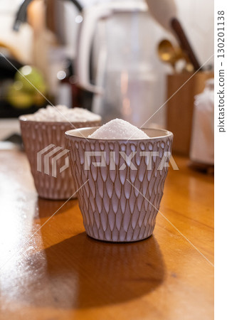 A ceramic cup filled with white sugar placed on a wooden kitchen counter, with another cup of flour blurred in the background 130201138
