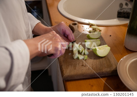A person slices green apples on a wooden cutting board next to a bowl of chopped apples and a cup of flour on the kitchen table 130201145