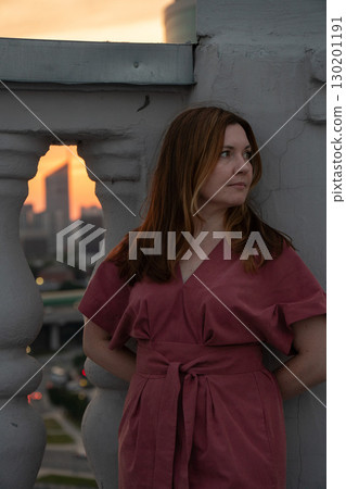 Woman in a rose dress leans against a rooftop balustrade at sunset with a blurred city skyline in the background 130201191