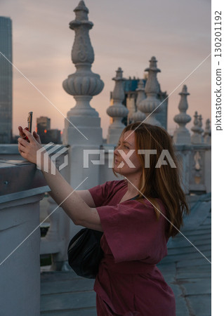 Woman in a rose dress takes a photo with her smartphone on a rooftop terrace at sunset, with ornate stone balustrades Woman in a rose dress takes a photo with her smartphone on a rooftop terrace at sunset, with ornate stone balustrades 130201192