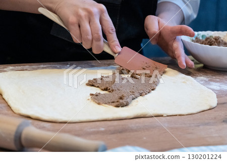 A woman spreads filling on dough with a spatula. A wooden table holds flour 130201224
