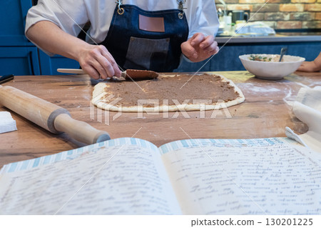 A woman spreads filling on dough with a spatula. An open cookbook with a handwritten recipe and a rolling pin are on the wooden table A woman spreads filling on dough with a spatula. An open cookbook with a handwritten recipe and a rolling pin are on the wooden table 130201225
