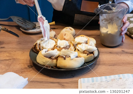 Woman spreading tasty cream onto baked cinnamon rolls on table in kitchen Woman spreading tasty cream onto baked cinnamon rolls on table in kitchen 130201247
