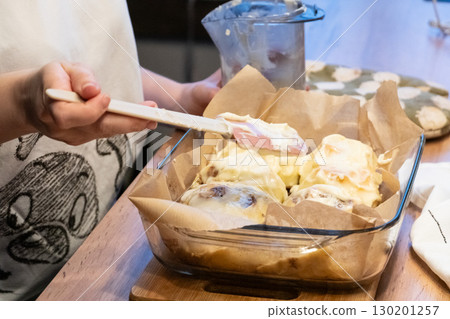 Woman spreading tasty cream onto baked cinnamon rolls on table in kitchen Woman spreading tasty cream onto baked cinnamon rolls on table in kitchen 130201257