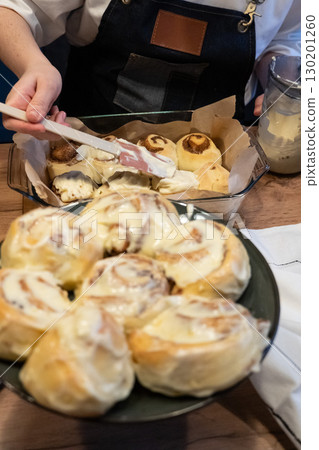 Woman spreading tasty cream onto baked cinnamon rolls on table in kitchen 130201260