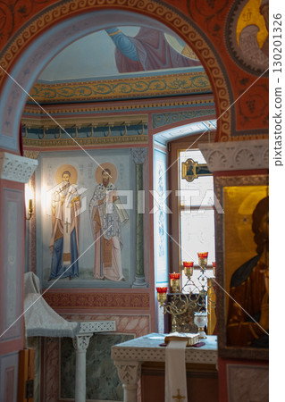 Interior of an Eastern Orthodox church with vivid iconography, frescoes of saints, and a decorated altar with red candle holders and a gold cross 130201326