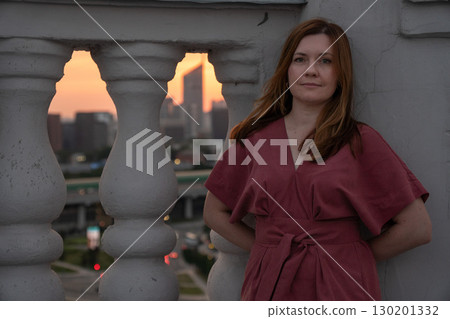 Woman in a rose dress leans against a rooftop balustrade at sunset with a blurred city skyline in the background Woman in a rose dress leans against a rooftop balustrade at sunset with a blurred city skyline in the background 130201332