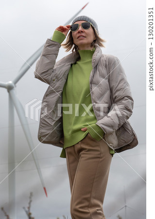 Woman in a beige puffer jacket and gray beanie adjusts her sunglasses with a wind turbine softly blurred in the background Woman in a beige puffer jacket and gray beanie adjusts her sunglasses with a wind turbine softly blurred in the background 130201351