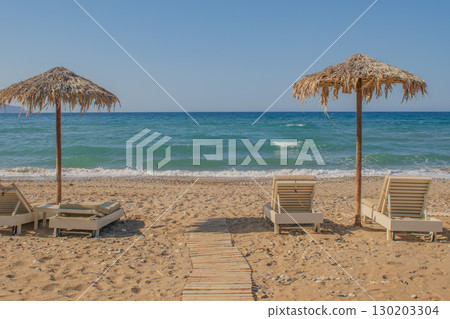 Simple Cretan beach scene with rustic umbrellas and sunbeds on a pebble shore Simple Cretan beach scene with rustic umbrellas and sunbeds on a pebble shore 130203304