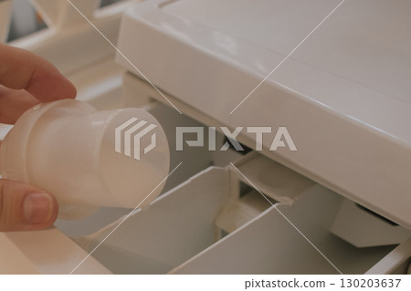 Female hand pouring fabric softener from cap into washing machine detergent compartment 130203637