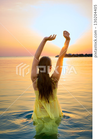 A young charming woman in a yellow dress stands on the seashore in front of a bright orange sunset. 130204405