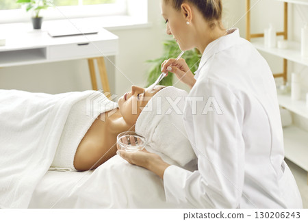Female cosmetologist applying moisturizing mask with brush to a young woman in spa salon. Female cosmetologist applying moisturizing mask with brush to a young woman in spa salon. 130206243